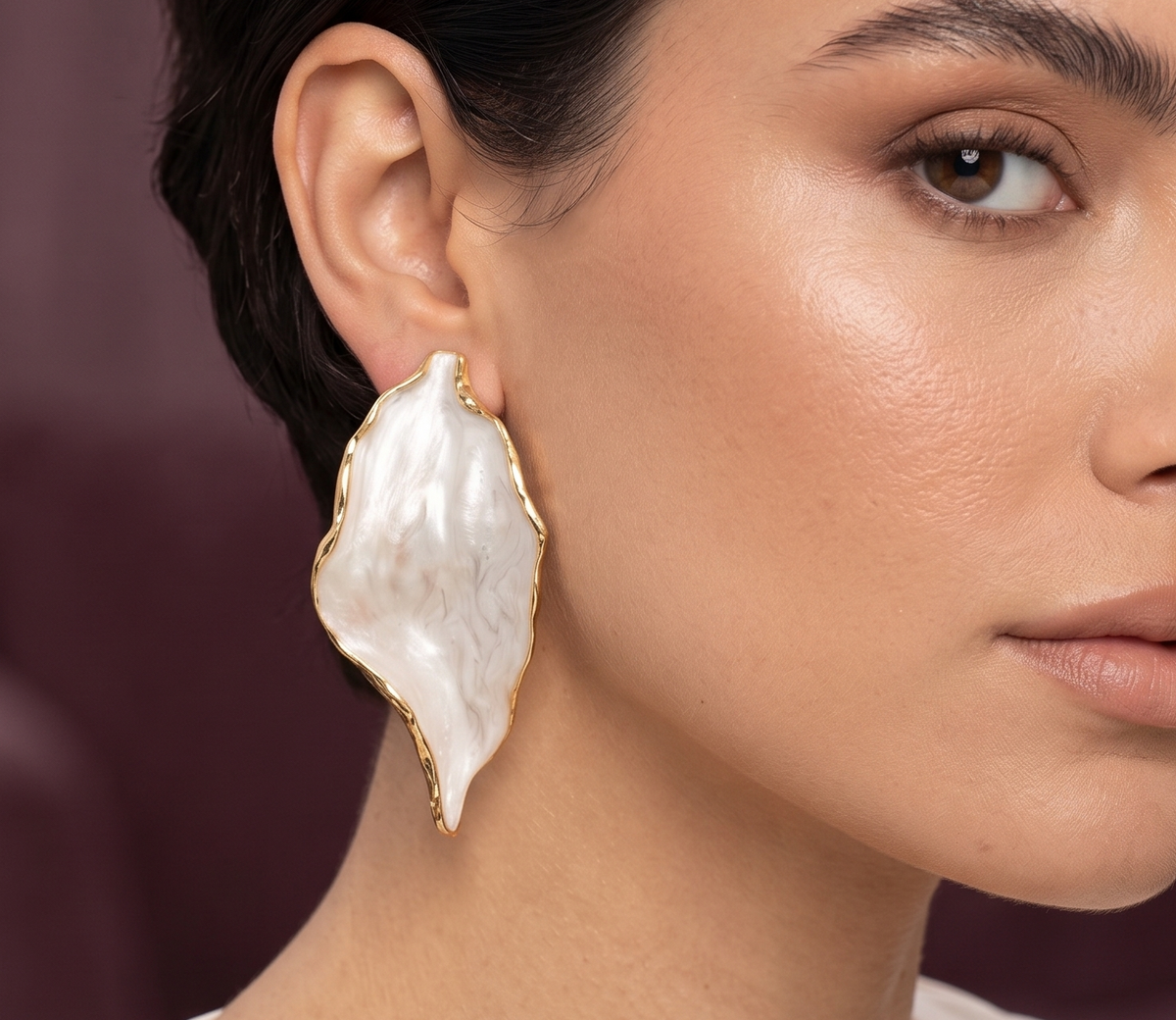 A close-up portrait of a woman wearing a large, organic-shaped white pearl earring with a gold-toned border, set against a soft, dark plum background.
